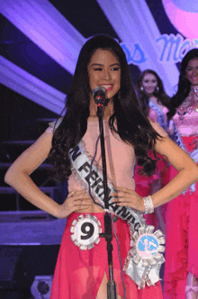 a woman stands in front of a microphone wearing a sash with the number 9