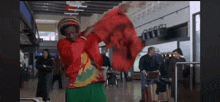 a man in a red jacket and green pants is holding a red scarf in an airport