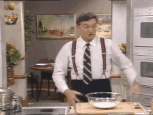 a man wearing suspenders and a tie is standing in a kitchen with a bowl of food on a cutting board