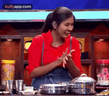 a woman in a red shirt and blue apron is standing in front of a table with pots and pans .