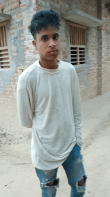 a young man in a white shirt and ripped jeans stands in front of a brick wall