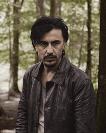 a man in a leather jacket stands in the woods looking at the camera