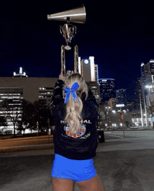 a cheerleader holds a trophy and a megaphone in front of a city skyline