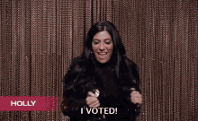 a woman is standing in front of a brown curtain and holding a button that says `` i voted '' .