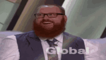 a man with a beard and glasses is sitting on a couch with the word global behind him .