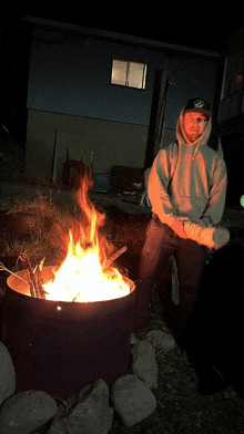 a man in a hoodie stands next to a fire in a barrel