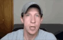 a man wearing a baseball cap and a gray shirt is sitting on a couch and talking to the camera .