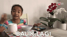 a little girl sits at a table with a bowl and a cup and the words balik lagi on the table cloth