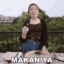 a woman is sitting at a table with a plate of food and the word makan ya written on it .