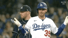 two baseball players are standing next to each other on a field . one of the players is wearing a dodgers jersey .