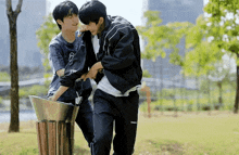 two young men are standing next to each other in a park . one of the men is holding an umbrella .