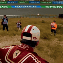 a man in a red ferrari jacket stands in a field with aramco banners behind him