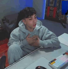 a man in a grey hoodie is sitting at a desk with his hands on his chest in front of a keyboard