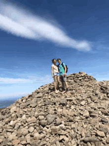 two people standing on top of a pile of rocks with a blue sky in the background