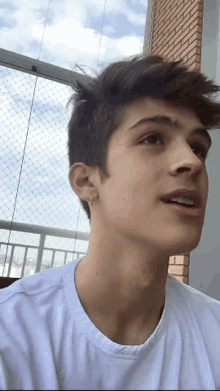 a young man wearing a white shirt and earrings looks at the camera with a brick building in the background