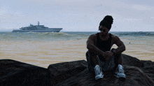 a man sits on a rock on a beach with a large ship in the background