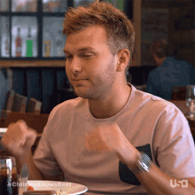 a man in a pink shirt is sitting at a table with a plate of food and a knife and fork