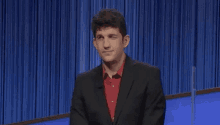 a man in a suit and red shirt is sitting on a stage with a blue curtain in the background .