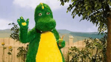 a green and yellow dinosaur mascot is standing in front of a fence .