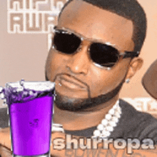 a man wearing sunglasses and a necklace is holding a purple drink