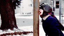 a young boy wearing a helmet and scarf is sticking his head through a chain link fence in the snow