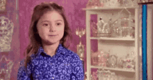 a little girl in a blue shirt is standing in front of a shelf filled with crowns and tiaras .