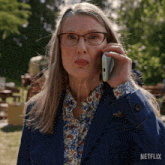 a woman wearing glasses and a denim jacket is talking on a cell phone with a netflix logo in the corner