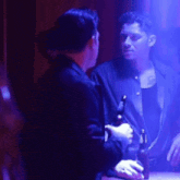 a man is holding a bottle of beer and talking to another man in a dark room
