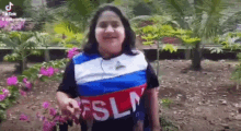 a woman wearing a blue , white and red shirt with the word fsln on it is standing in front of flowers .