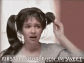 a woman with pigtails is holding her hair in a bun and says `` first im sour then im sweet '' .
