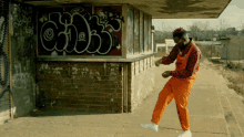 a man in orange overalls is dancing in front of a brick building with graffiti on it that says ' a '