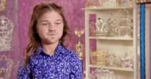 a young man with long hair is wearing a blue shirt and standing in front of a shelf full of crowns .