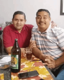 two men are sitting at a table with a bottle of beer and a mug of beer .