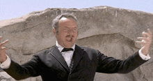 a man in a tuxedo and bow tie is standing in front of a rock .