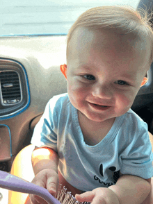 a baby wearing a blue shirt with a mickey mouse on it is smiling while holding a purple brush