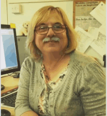 a woman with glasses and a mustache is smiling in front of a wall with a bulletin board that says bus helps