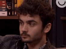 a young man with curly hair and a beard is sitting in front of a bookshelf with his eyes closed .