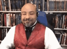 a bald man with a beard is wearing a red vest and bow tie while sitting in front of a bookshelf .