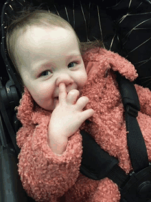 a baby wearing a pink jacket is sitting in a car seat with his finger in his nose