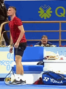 a man in a red shirt is standing on a tennis court with a yonex bag in front of him