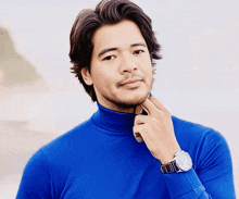 a man wearing a blue turtleneck and a watch holds his hand to his chin