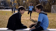 two boys are sitting on a bench talking to each other while a girl in a blue shirt runs in the background