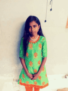 a young girl in a green and orange dress stands in front of a wall