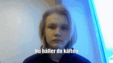 a young man with long blonde hair is standing in front of a window with the words nu håller du kaften above him