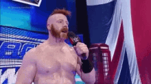 a shirtless wrestler with a beard is holding a microphone in front of a british flag .