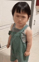 a little girl wearing green overalls and a grey tank top is making a funny face