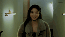 a woman wearing a fur coat is smiling in a dark room with a light on the wall behind her