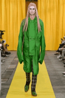 a man with long blonde hair is walking down the runway at a fashion show wearing a green coat and plaid pants .