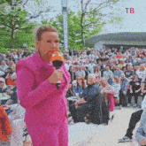 a woman in a pink suit holds a microphone in front of a crowd with the letters tb on the bottom right