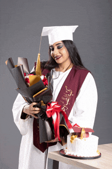 a woman in a graduation cap and gown holds a bouquet of flowers and a cake